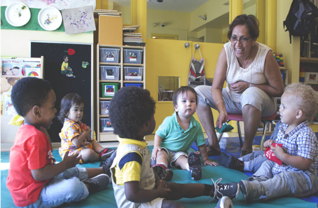 A childcare teacher sitting on a chair leads a singing circle with five toddlers sitting on a green mat.